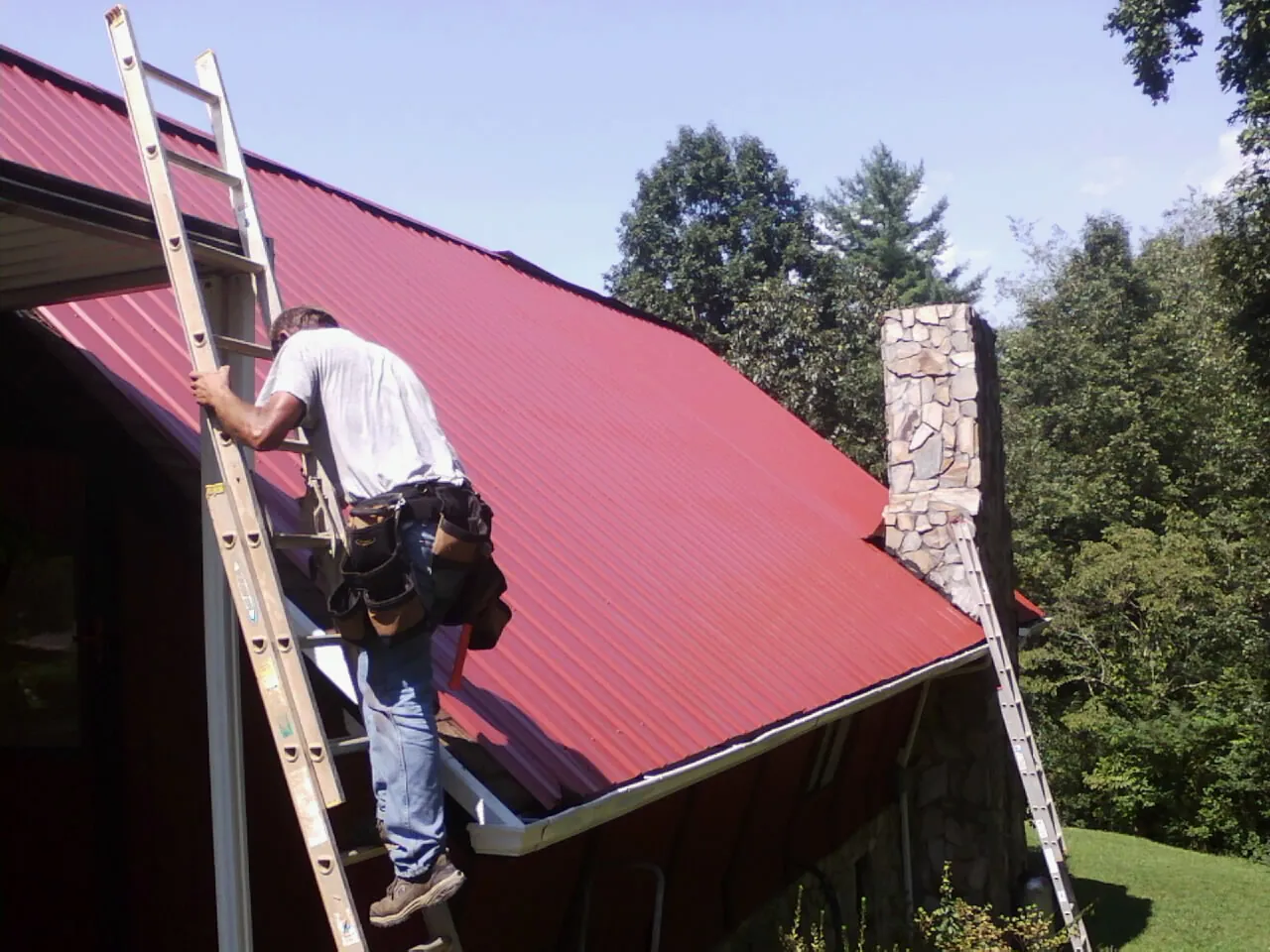 Roofer on a ladder beside a red metal roof and stone chimney — Soffit & Fascia Repair by Roofing Brothers in La Rue